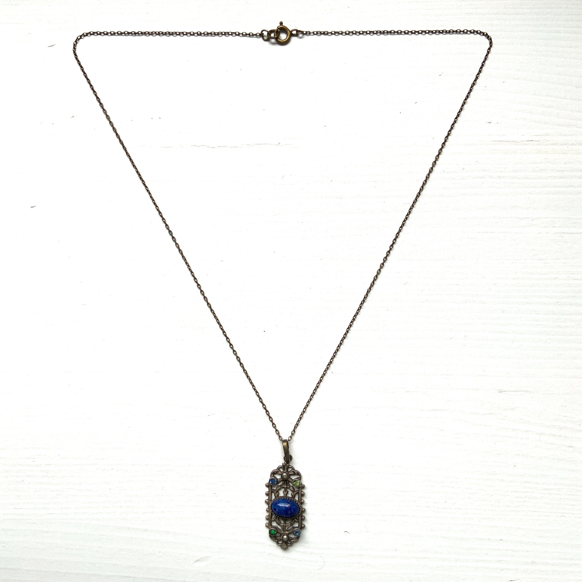 A delicate filigree pendant necklace adorned with a lapis glass cabochon, colourful rhinestones, and seed glass pearls. 繊細なフィリグリーのプレートに、ラピスグラスのカボションとカラフルなラインストーン、シードグラスパールがあしらわれたペンダントネックレス。