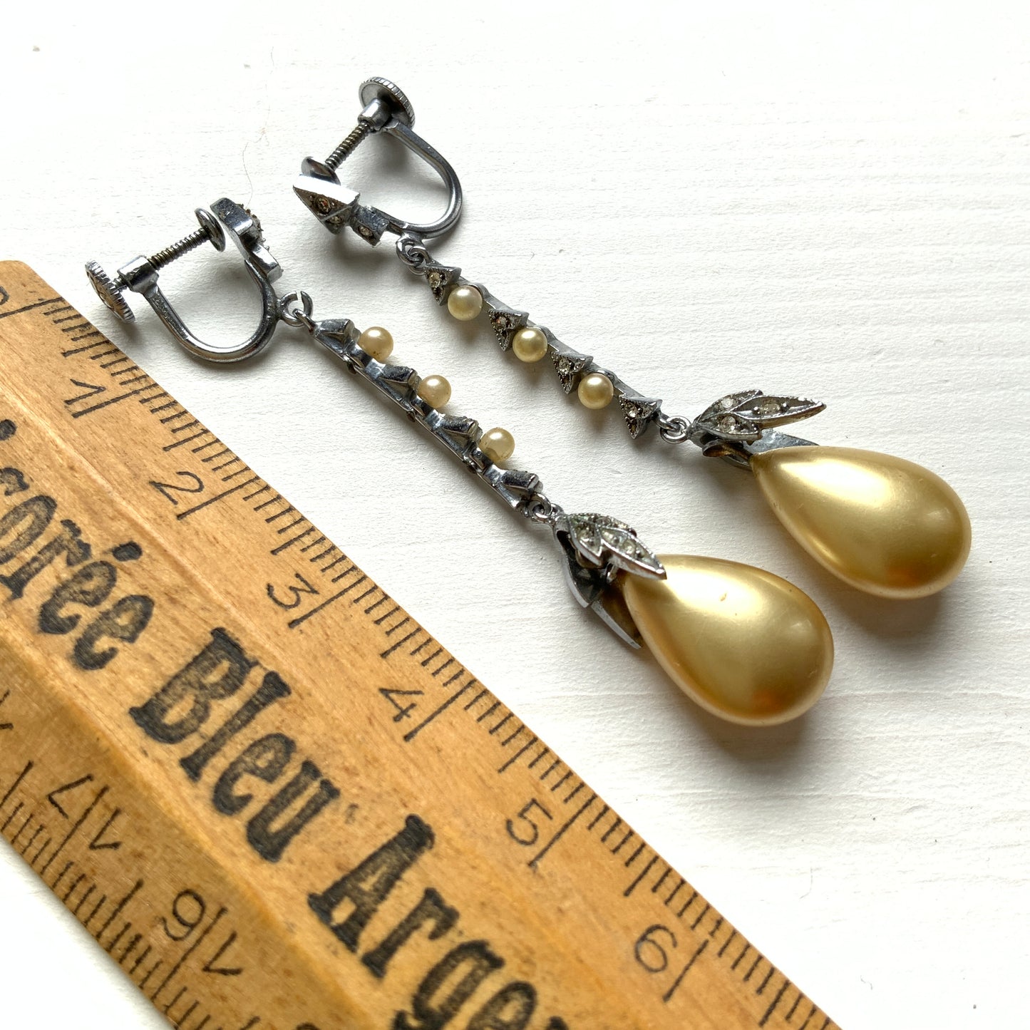 Screw earrings featuring a silver-tone leaf with a champagne gold teardrop pearl glass dangling from the tip. シルバートーンのリーフの先にシャンパンゴールドカラーのティアドロップグラスパールが揺れるスクリューイヤリング。