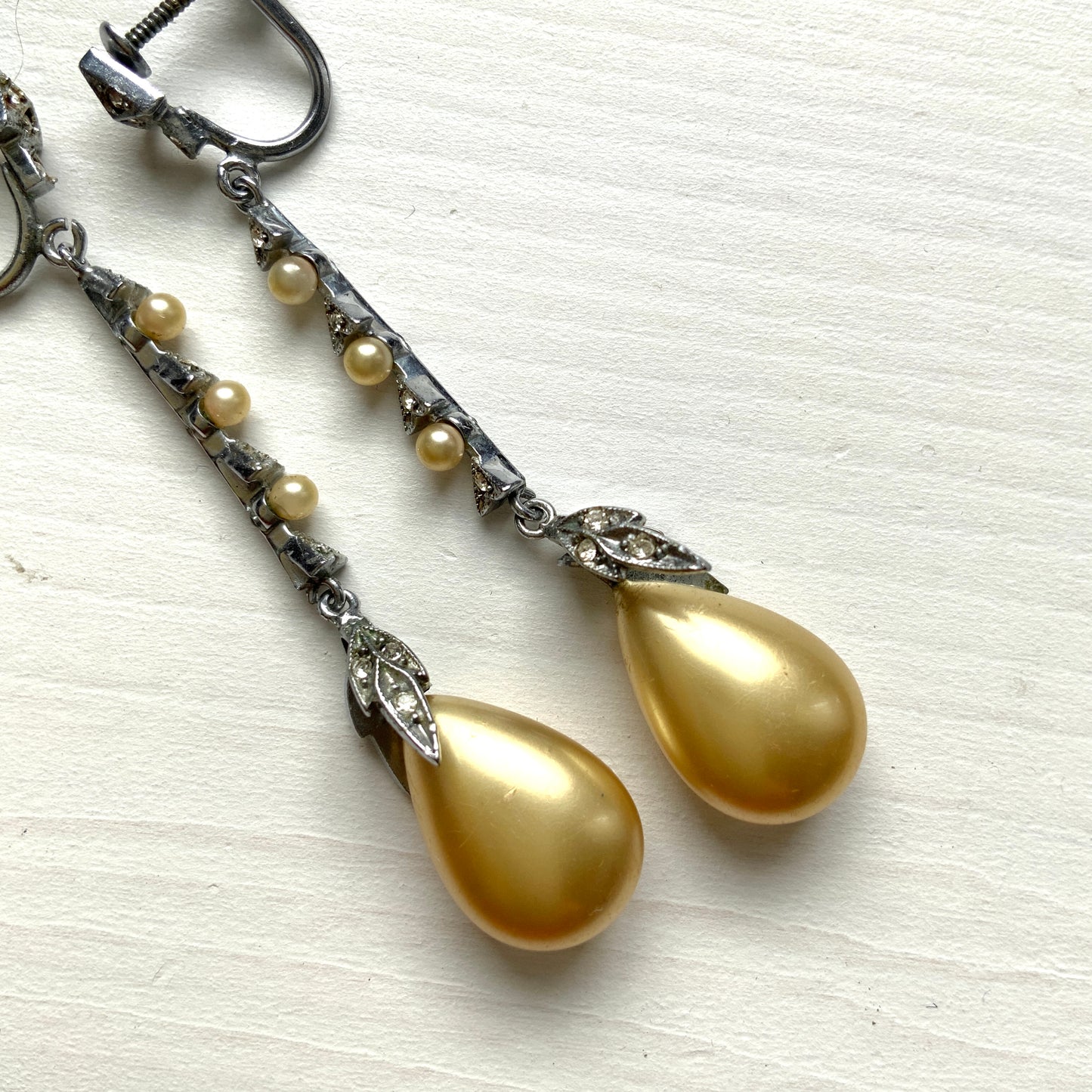 Screw earrings featuring a silver-tone leaf with a champagne gold teardrop pearl glass dangling from the tip. シルバートーンのリーフの先にシャンパンゴールドカラーのティアドロップグラスパールが揺れるスクリューイヤリング。