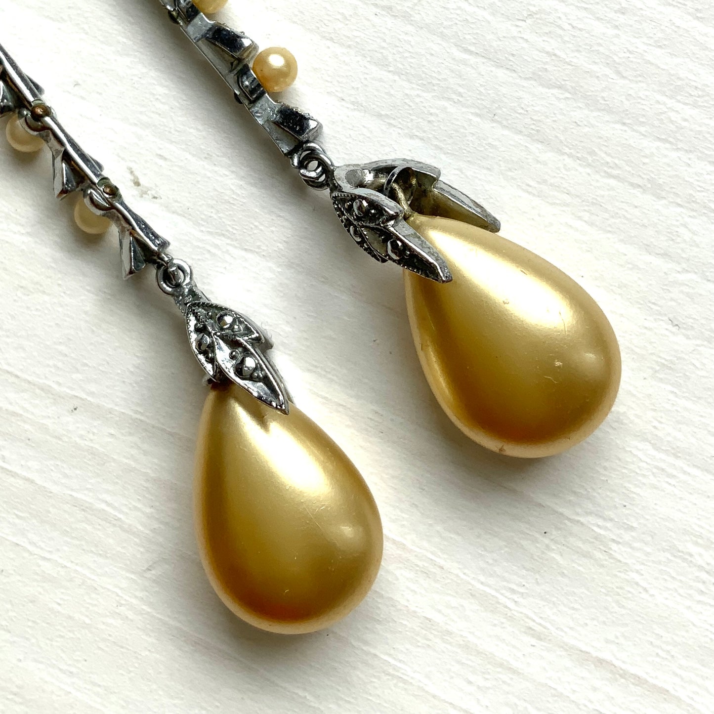 Screw earrings featuring a silver-tone leaf with a champagne gold teardrop pearl glass dangling from the tip. シルバートーンのリーフの先にシャンパンゴールドカラーのティアドロップグラスパールが揺れるスクリューイヤリング。