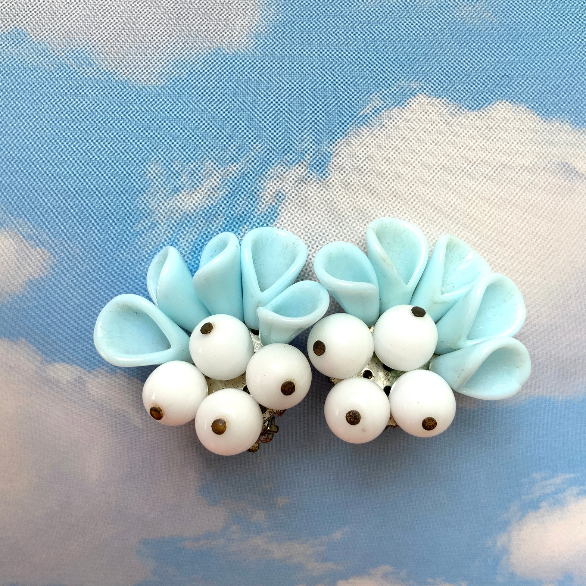 These clip-on earrings feature petal-like pastel blue glass beads paired with white milk glass beads that resemble berries. 花びらのような淡いパステルブルーのグラスビーズに、白い実のようなミルクグラスビーズがついたクリップイヤリング。