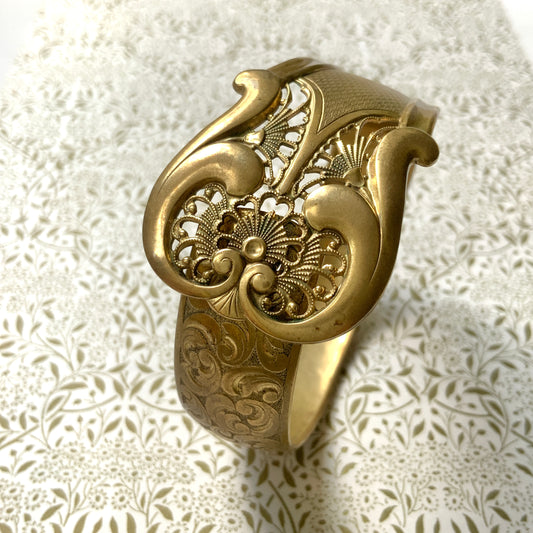 This gold-tone metal bangle features an elegant buckle adorned with delicate filigree work.  フィリグリーで装飾されたエレガントなバックルが印象的なゴールドトーンメタルのバングル。