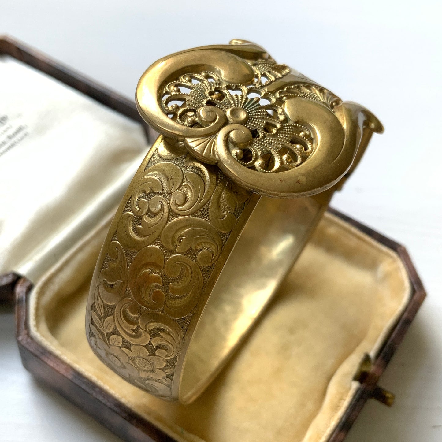 This gold-tone metal bangle features an elegant buckle adorned with delicate filigree work.  フィリグリーで装飾されたエレガントなバックルが印象的なゴールドトーンメタルのバングル。