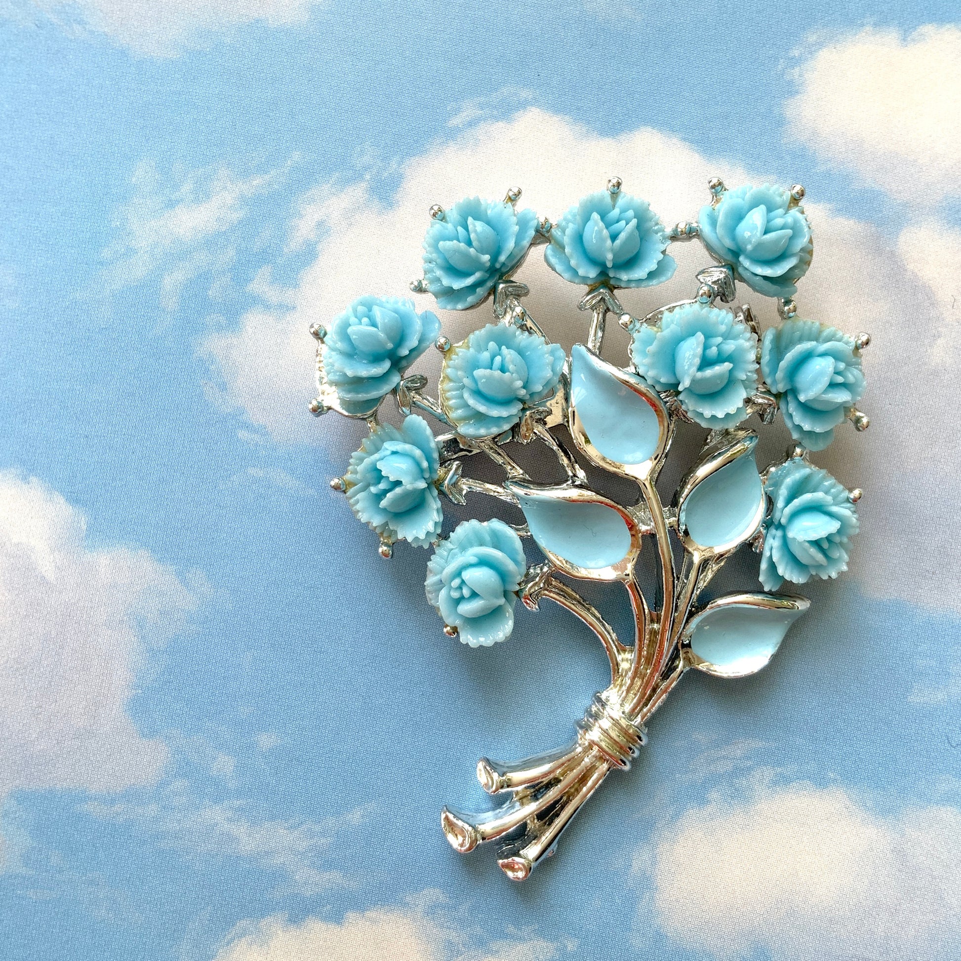 A bouquet brooch featuring sky blue plastic roses, with matching enamel-coated leaves and silver-tone branches that beautifully complement the color scheme. 空色のプラスチックのバラのブーケブローチ。