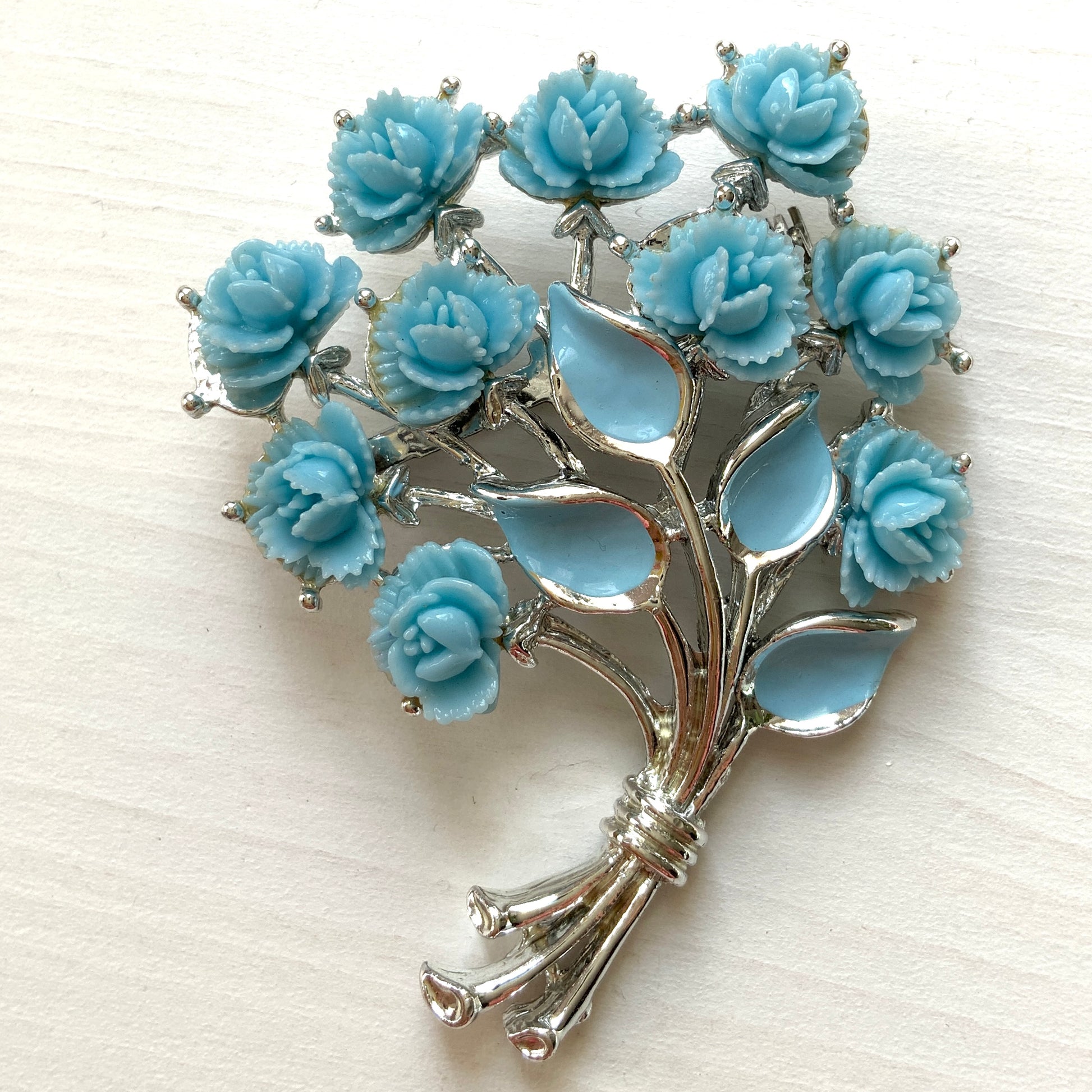 A bouquet brooch featuring sky blue plastic roses, with matching enamel-coated leaves and silver-tone branches that beautifully complement the color scheme. 空色のプラスチックのバラのブーケブローチ。