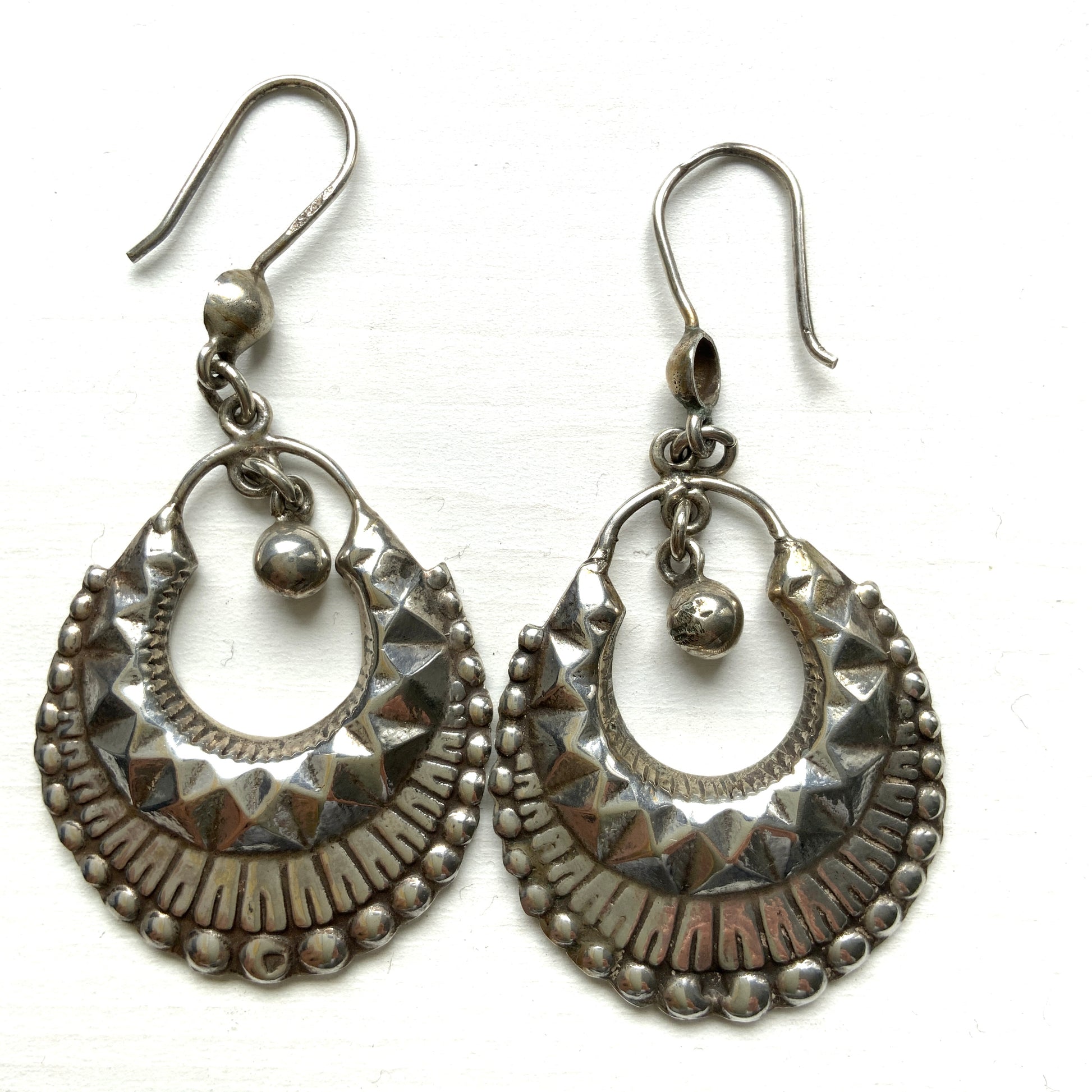 Striking silver tone hoop earrings with geometric embossed patterns and granulated bead details. 半月型にジオメトリックな打ち出し模様と粒状の装飾を施した、存在感あるシルバートーンのフープピアス。