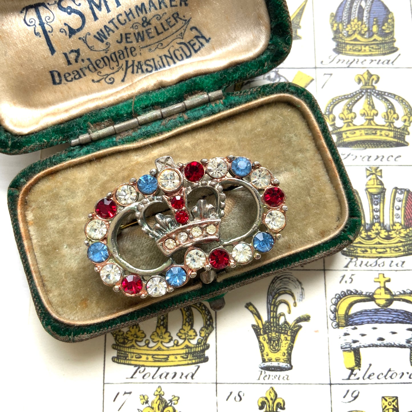 A brooch featuring a crown encircled by tricolour rhinestones – red, clear and blue. 王冠の周りをトリコロールカラーのラインストーンが囲んだブローチ。