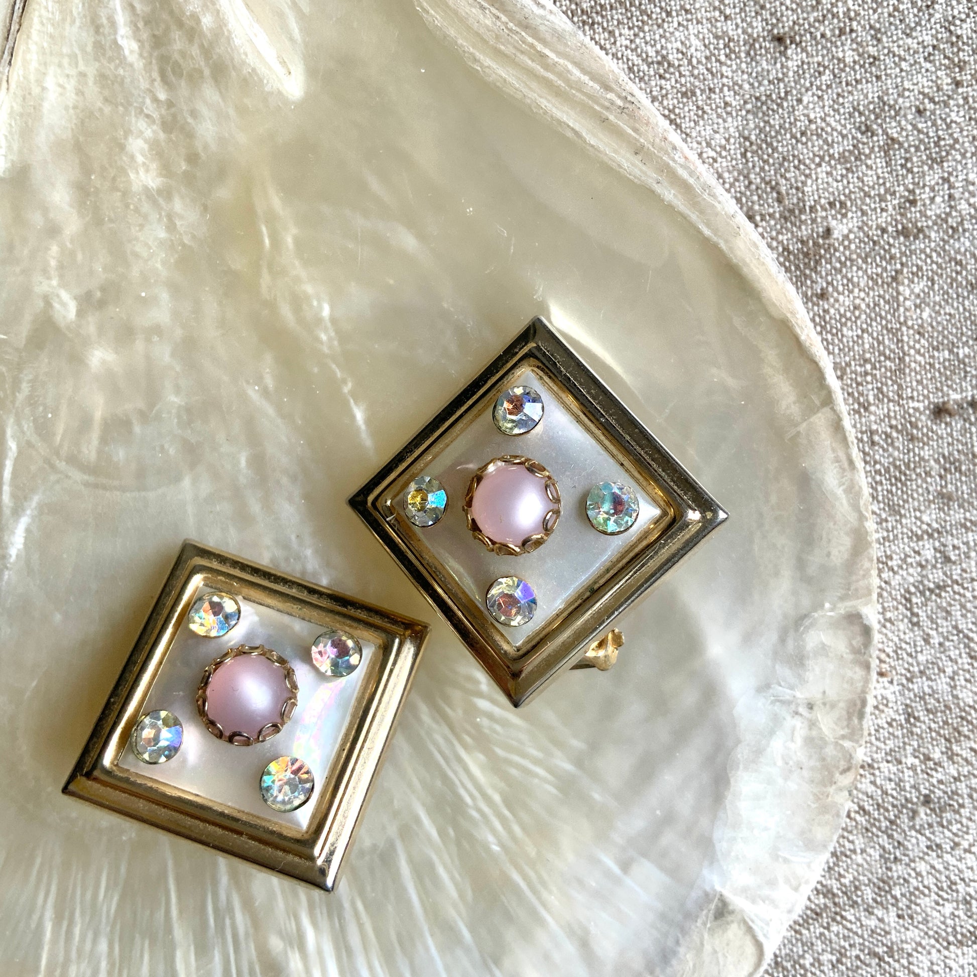 Clip-on earrings featuring white square plastic made to resemble mother of pearl, accented with pink plastic cabochons and shimmering aurora borealis stones. マザーオブパールを模したホワイトのスクエアプラスチックに、ピンクのプラスチックカボションとオーロラストーンが輝くクリップイヤリング。