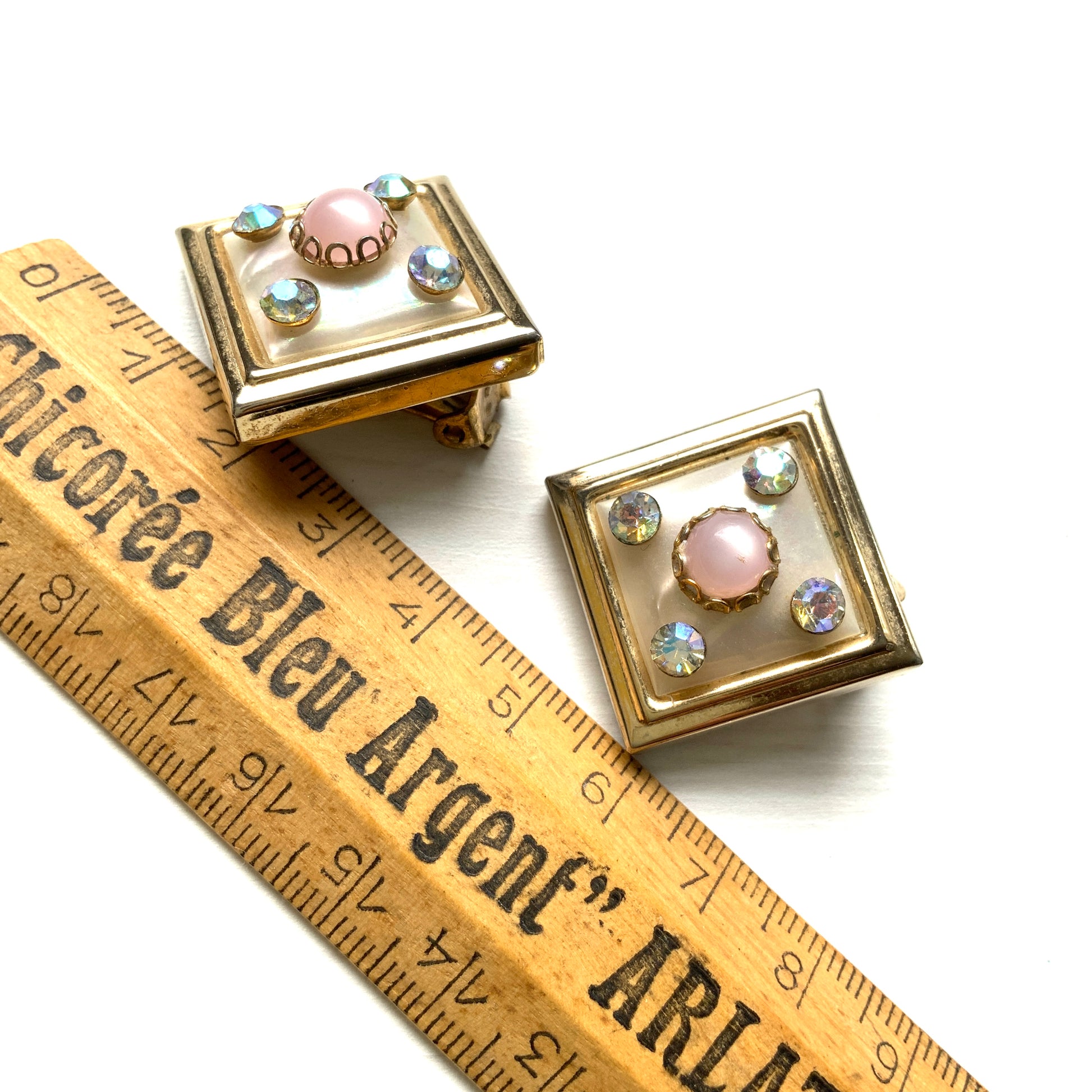 Clip-on earrings featuring white square plastic made to resemble mother of pearl, accented with pink plastic cabochons and shimmering aurora borealis stones. マザーオブパールを模したホワイトのスクエアプラスチックに、ピンクのプラスチックカボションとオーロラストーンが輝くクリップイヤリング。