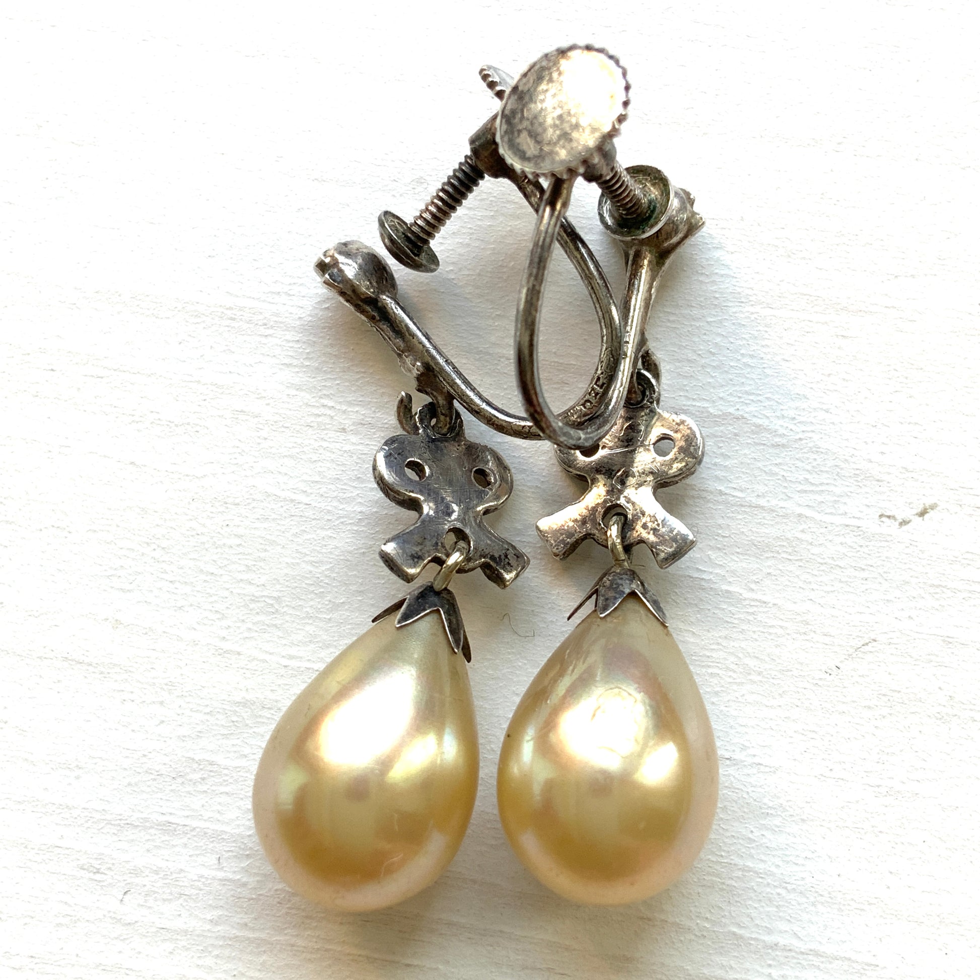 A pair of screw-back earrings featuring teardrop glass pearls suspended from silver bows. シルバー製のリボンに、ティアドロップのグラスパールが揺れるスクリューイヤリング。
