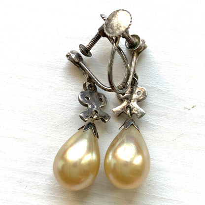 A pair of screw-back earrings featuring teardrop glass pearls suspended from silver bows. シルバー製のリボンに、ティアドロップのグラスパールが揺れるスクリューイヤリング。