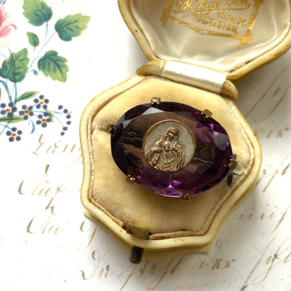 An antique brooch featuring a medal of the Virgin Mary set at the centre of a deep purple faceted glass stone. 深いパープルのカットガラスの中央に、聖母マリアを象ったメダルをセットしたアンティークブローチ。