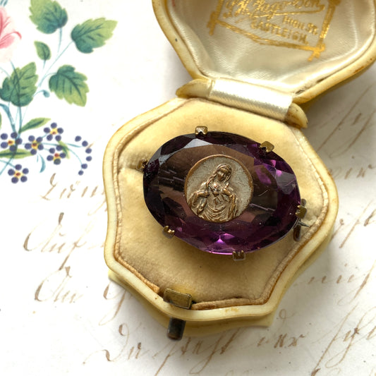 An antique brooch featuring a medal of the Virgin Mary set at the centre of a deep purple faceted glass stone. 深いパープルのカットガラスの中央に、聖母マリアを象ったメダルをセットしたアンティークブローチ。