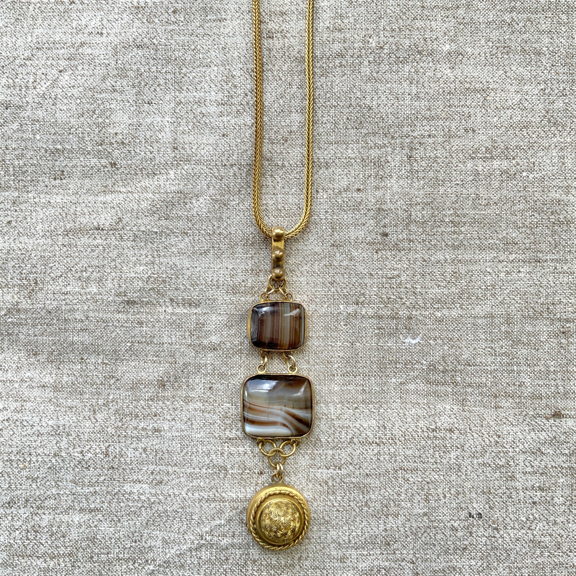 A lavaliere necklace featuring beautifully banded brown and white agate, paired with a gold-tone charm. ブラウンとホワイトの縞模様が美しいメノウと、ゴールドトーンのチャームが組み合わされたラヴァリエネックレス。