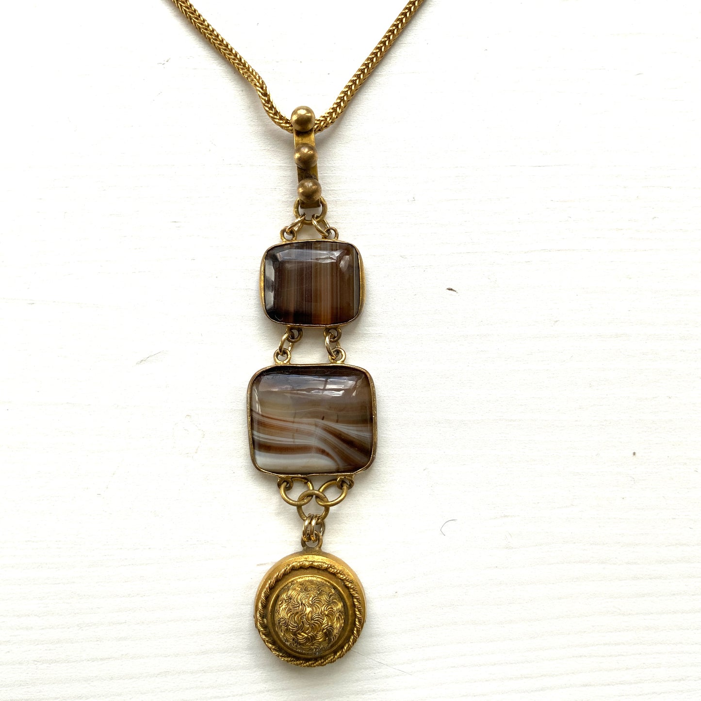 A lavaliere necklace featuring beautifully banded brown and white agate, paired with a gold-tone charm. ブラウンとホワイトの縞模様が美しいメノウと、ゴールドトーンのチャームが組み合わされたラヴァリエネックレス。