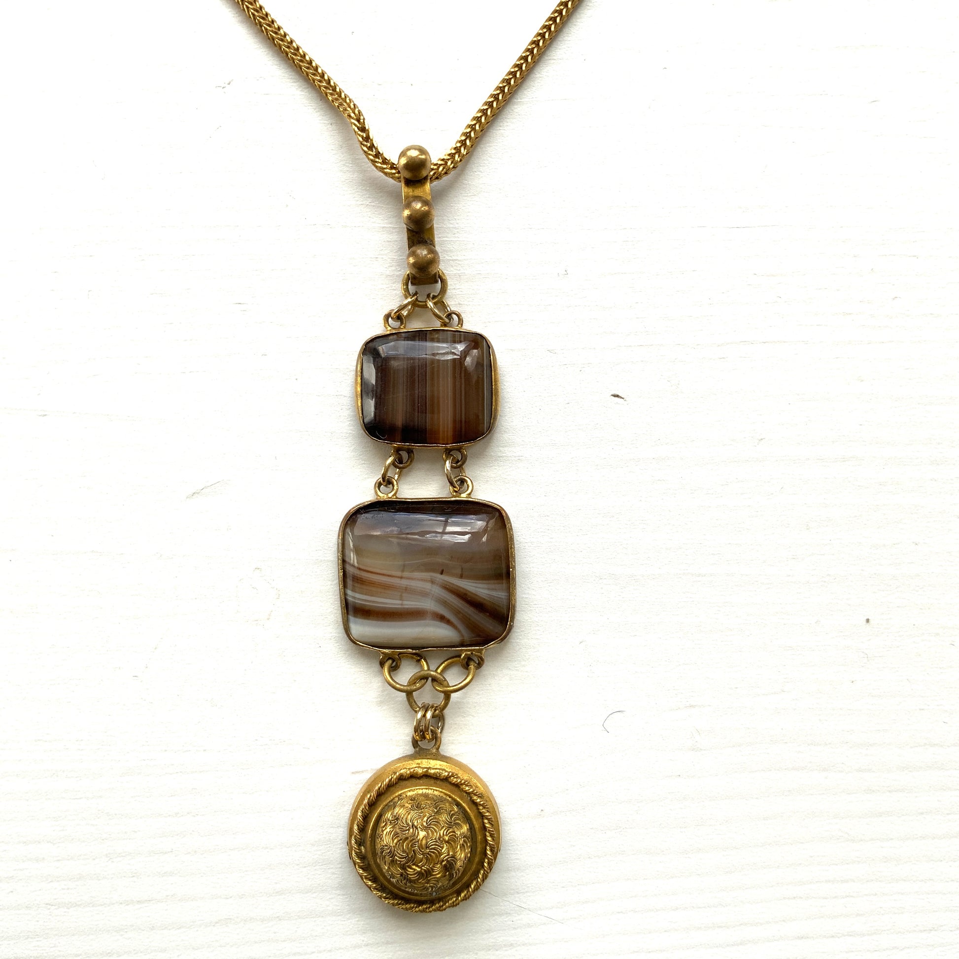 A lavaliere necklace featuring beautifully banded brown and white agate, paired with a gold-tone charm. ブラウンとホワイトの縞模様が美しいメノウと、ゴールドトーンのチャームが組み合わされたラヴァリエネックレス。