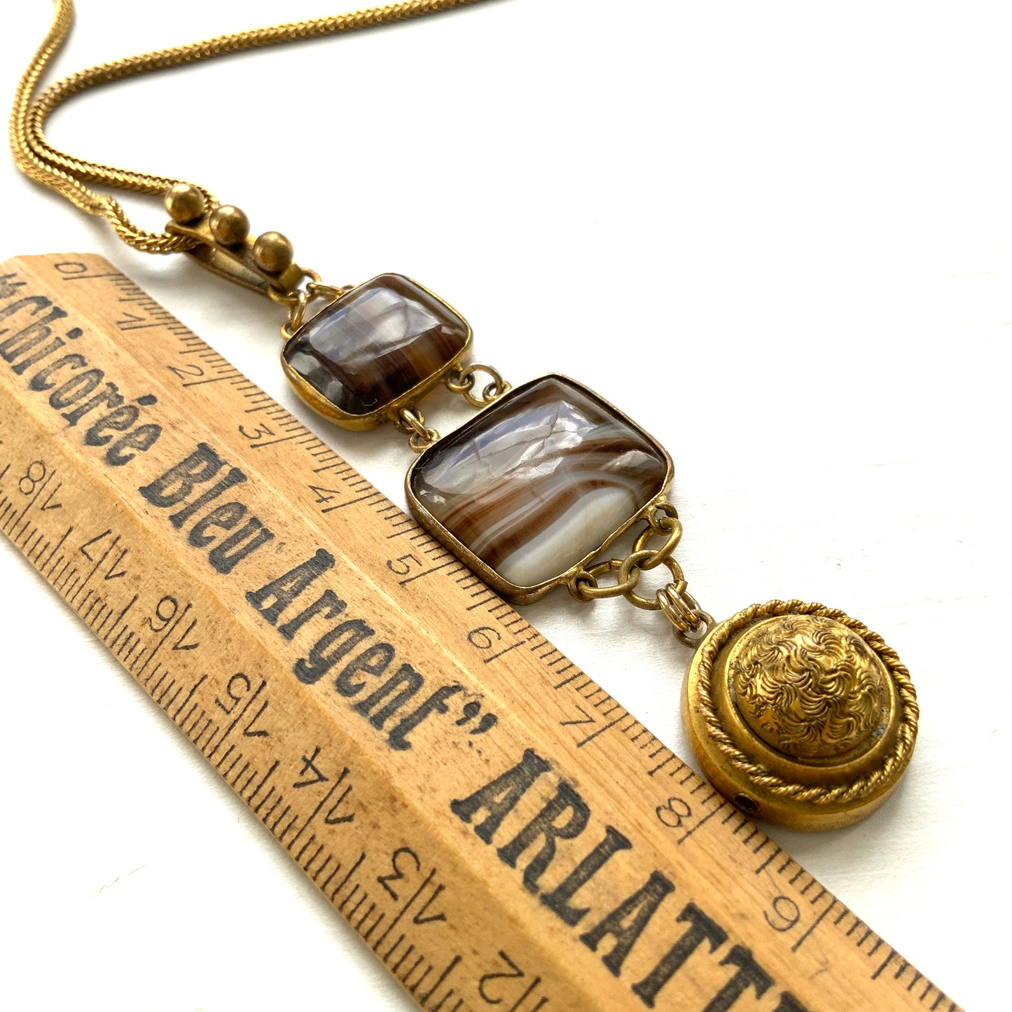 A lavaliere necklace featuring beautifully banded brown and white agate, paired with a gold-tone charm. ブラウンとホワイトの縞模様が美しいメノウと、ゴールドトーンのチャームが組み合わされたラヴァリエネックレス。