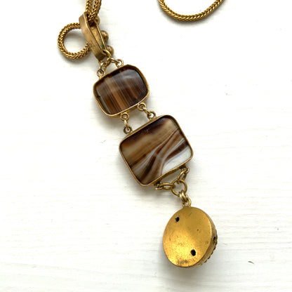 A lavaliere necklace featuring beautifully banded brown and white agate, paired with a gold-tone charm. ブラウンとホワイトの縞模様が美しいメノウと、ゴールドトーンのチャームが組み合わされたラヴァリエネックレス。
