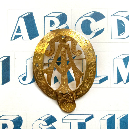 A gold-tone monogram brooch beautifully engraved with a delicate botanical motif.  美しい植物モチーフが彫られたゴールドトーンのモノグラムブローチ。