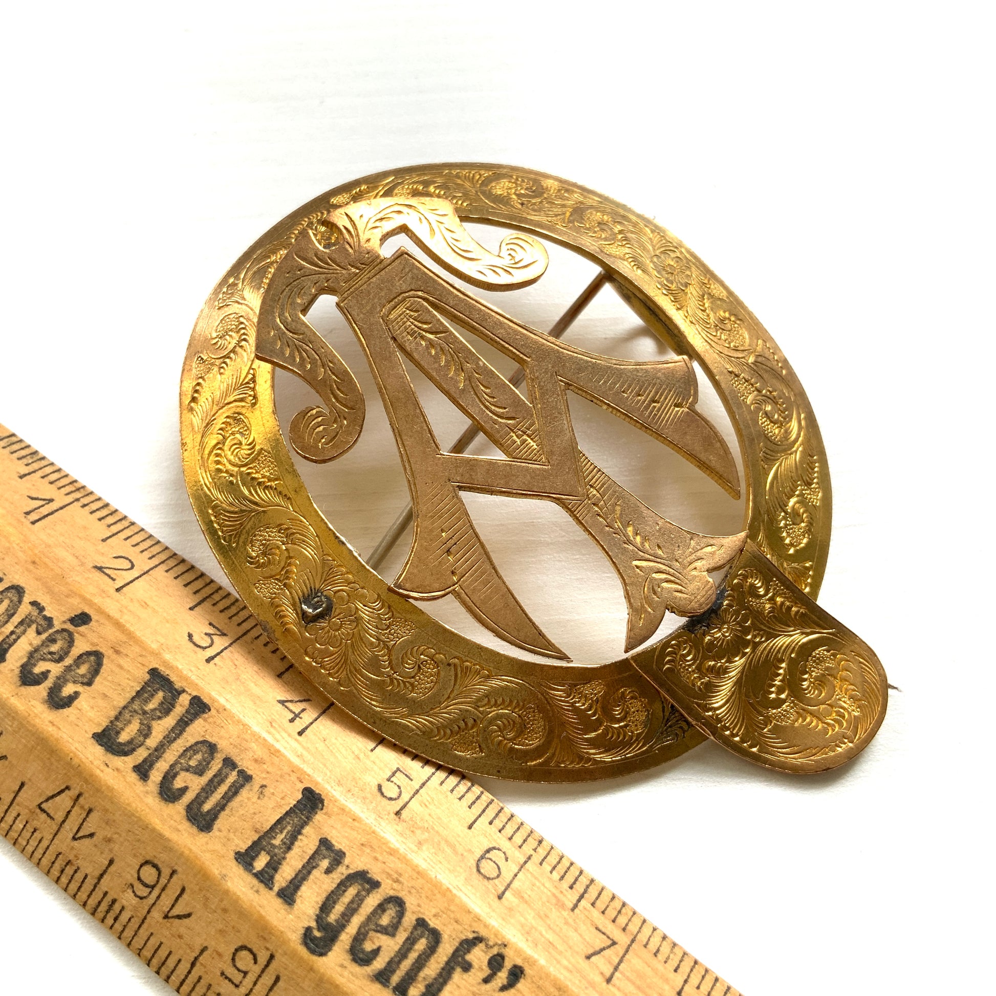 A gold-tone monogram brooch beautifully engraved with a delicate botanical motif.  美しい植物モチーフが彫られたゴールドトーンのモノグラムブローチ。