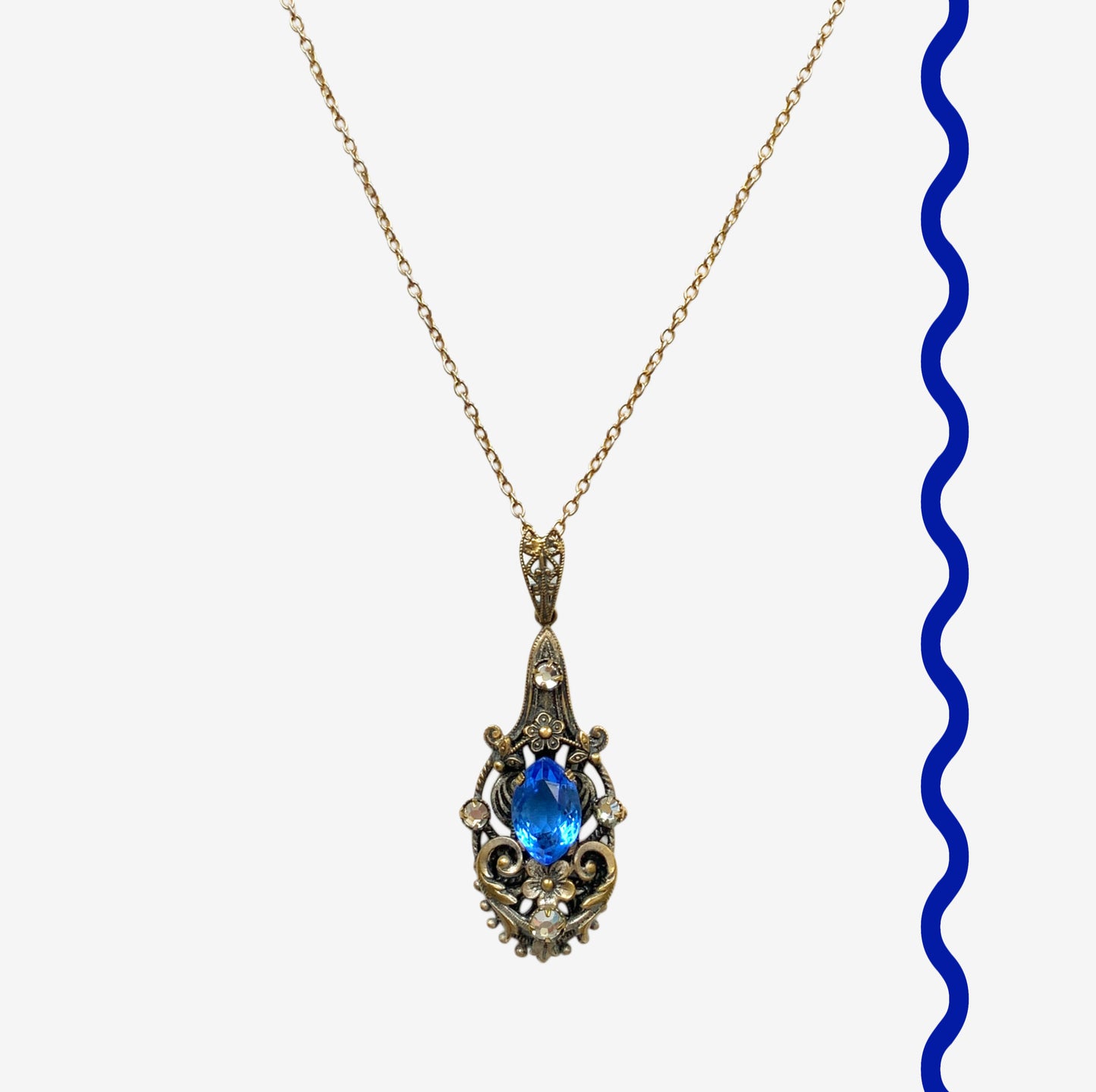 A vintage pendant necklace featuring a vivid blue navette glass stone surrounded by floral motifs. 鮮やかなブルーのナヴェットグラスの周りに、フローラルモチーフがあしらわれたヴィンテージのペンダントネックレス。