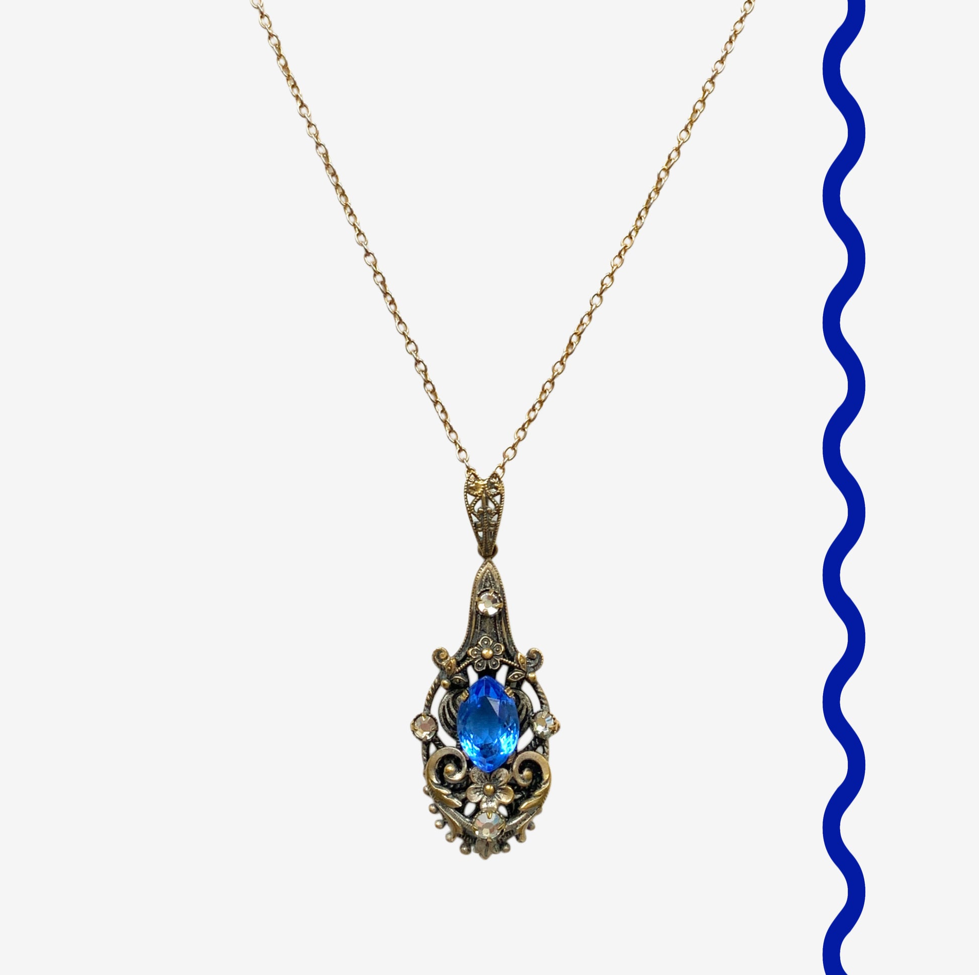 A vintage pendant necklace featuring a vivid blue navette glass stone surrounded by floral motifs. 鮮やかなブルーのナヴェットグラスの周りに、フローラルモチーフがあしらわれたヴィンテージのペンダントネックレス。