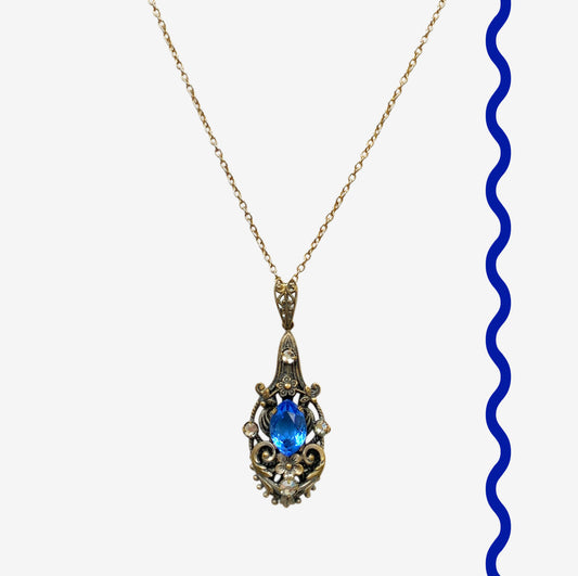 A vintage pendant necklace featuring a vivid blue navette glass stone surrounded by floral motifs. 鮮やかなブルーのナヴェットグラスの周りに、フローラルモチーフがあしらわれたヴィンテージのペンダントネックレス。