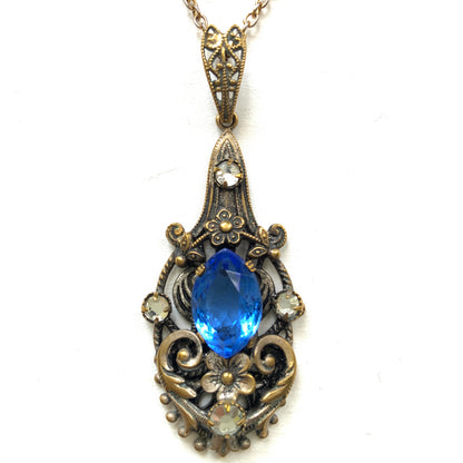 A vintage pendant necklace featuring a vivid blue navette glass stone surrounded by floral motifs. 鮮やかなブルーのナヴェットグラスの周りに、フローラルモチーフがあしらわれたヴィンテージのペンダントネックレス。