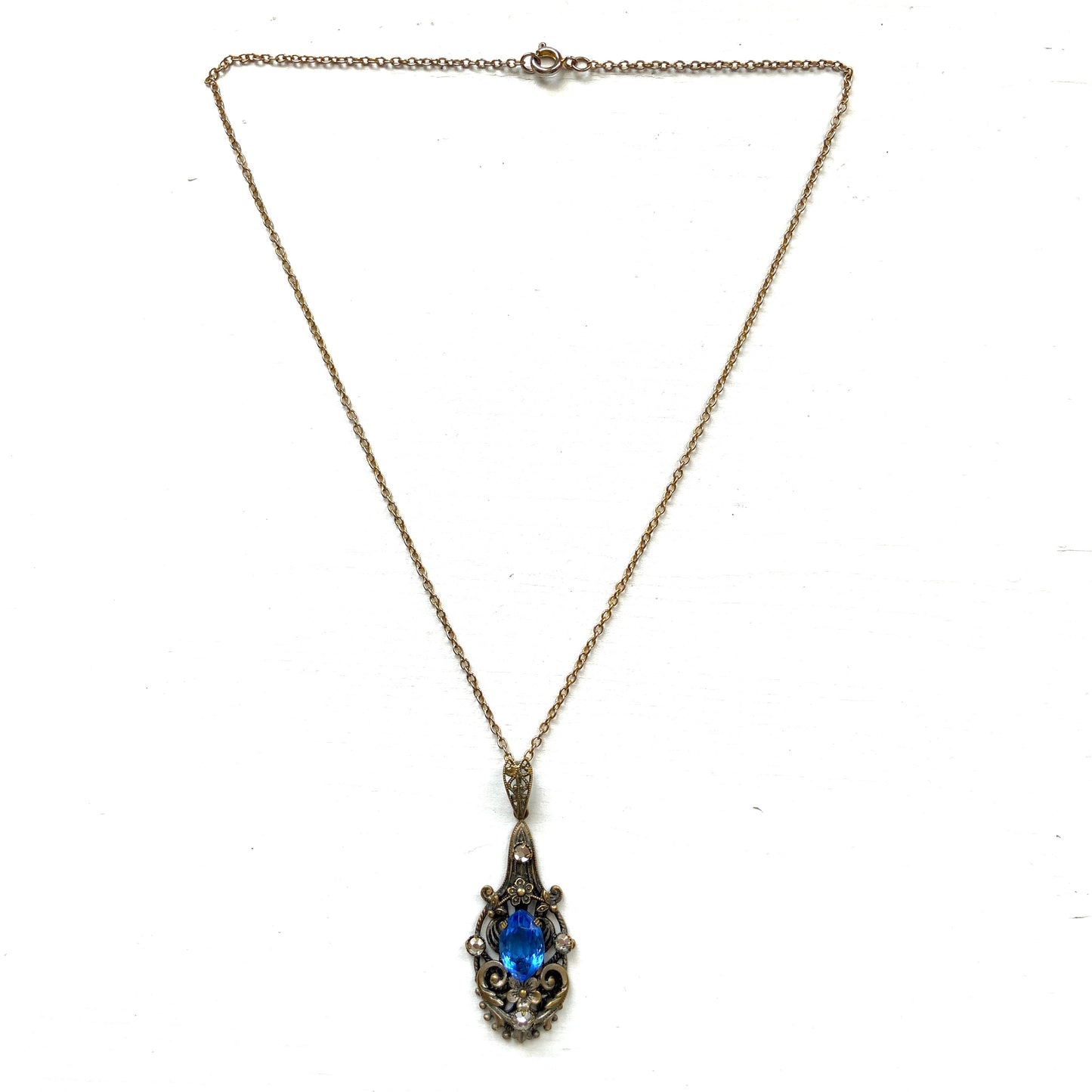 A vintage pendant necklace featuring a vivid blue navette glass stone surrounded by floral motifs. 鮮やかなブルーのナヴェットグラスの周りに、フローラルモチーフがあしらわれたヴィンテージのペンダントネックレス。
