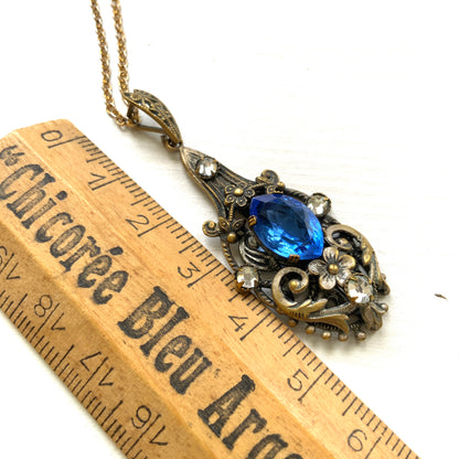 A vintage pendant necklace featuring a vivid blue navette glass stone surrounded by floral motifs. 鮮やかなブルーのナヴェットグラスの周りに、フローラルモチーフがあしらわれたヴィンテージのペンダントネックレス。