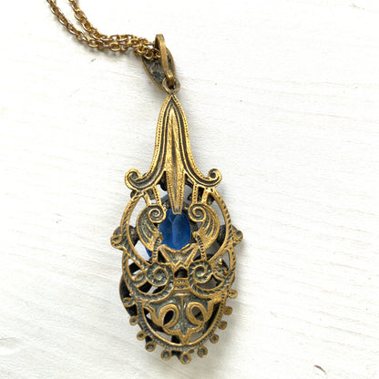 A vintage pendant necklace featuring a vivid blue navette glass stone surrounded by floral motifs. 鮮やかなブルーのナヴェットグラスの周りに、フローラルモチーフがあしらわれたヴィンテージのペンダントネックレス。