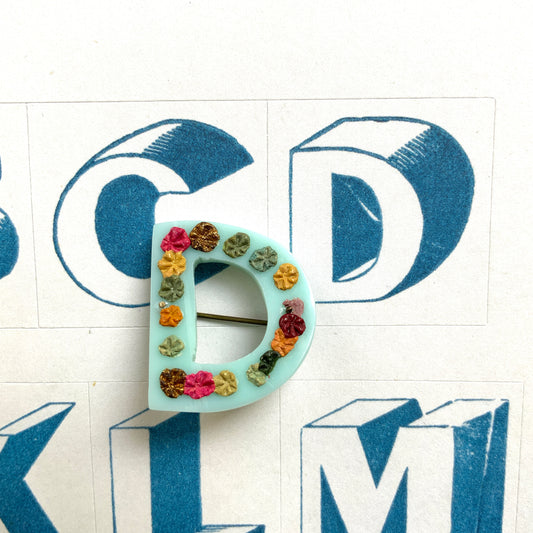 A brooch in the shape of the letter “D”, crafted from pale blue early plastic and adorned with colourful clay flowers. 淡い水色のアーリープラスチックに、カラフルなクレイのお花が咲いた”D"のブローチ。
