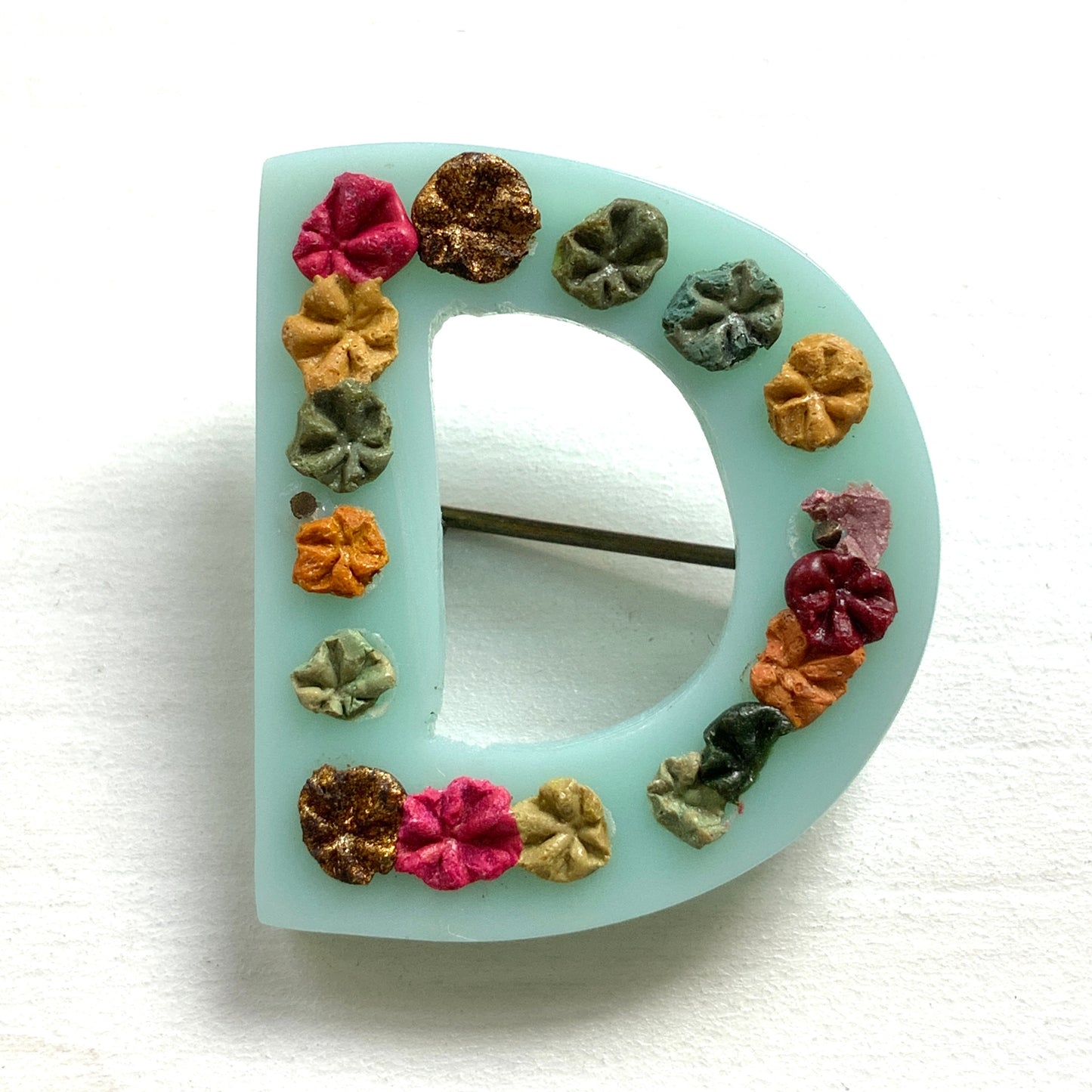 A brooch in the shape of the letter “D”, crafted from pale blue early plastic and adorned with colourful clay flowers. 淡い水色のアーリープラスチックに、カラフルなクレイのお花が咲いた”D"のブローチ。