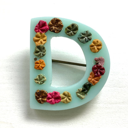 A brooch in the shape of the letter “D”, crafted from pale blue early plastic and adorned with colourful clay flowers. 淡い水色のアーリープラスチックに、カラフルなクレイのお花が咲いた”D"のブローチ。