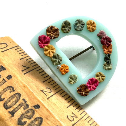 A brooch in the shape of the letter “D”, crafted from pale blue early plastic and adorned with colourful clay flowers. 淡い水色のアーリープラスチックに、カラフルなクレイのお花が咲いた”D"のブローチ。
