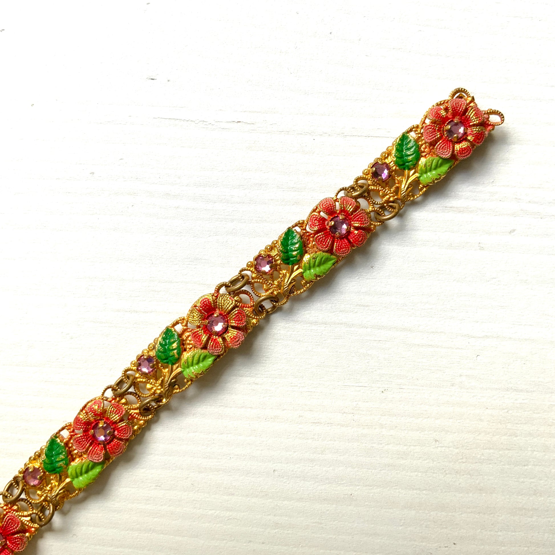 This gold-tone filigree bracelet features charming pink flowers delicately painted onto its dainty filigree plates. かわいらしいピンクのお花が描かれたゴールドトーンのフィリグリーのブレスレット。