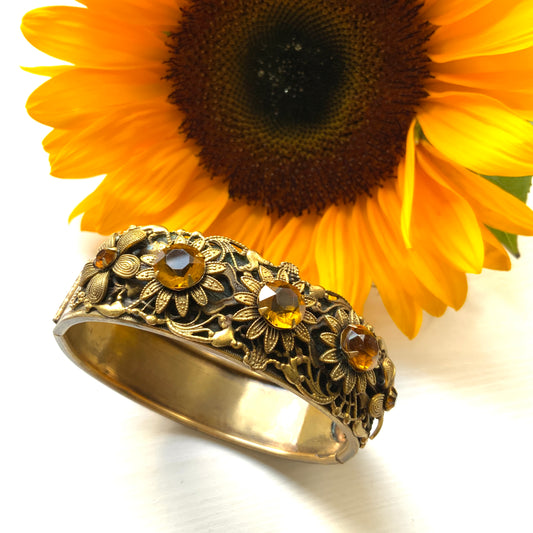 A decorative bangle featuring intricate floral motifs on gold-tone metal, with a filigree inspired design.  ゴールドトーンのメタルに、フィリグリー風の繊細な花模様があしらわれたデコラティブなバングル。