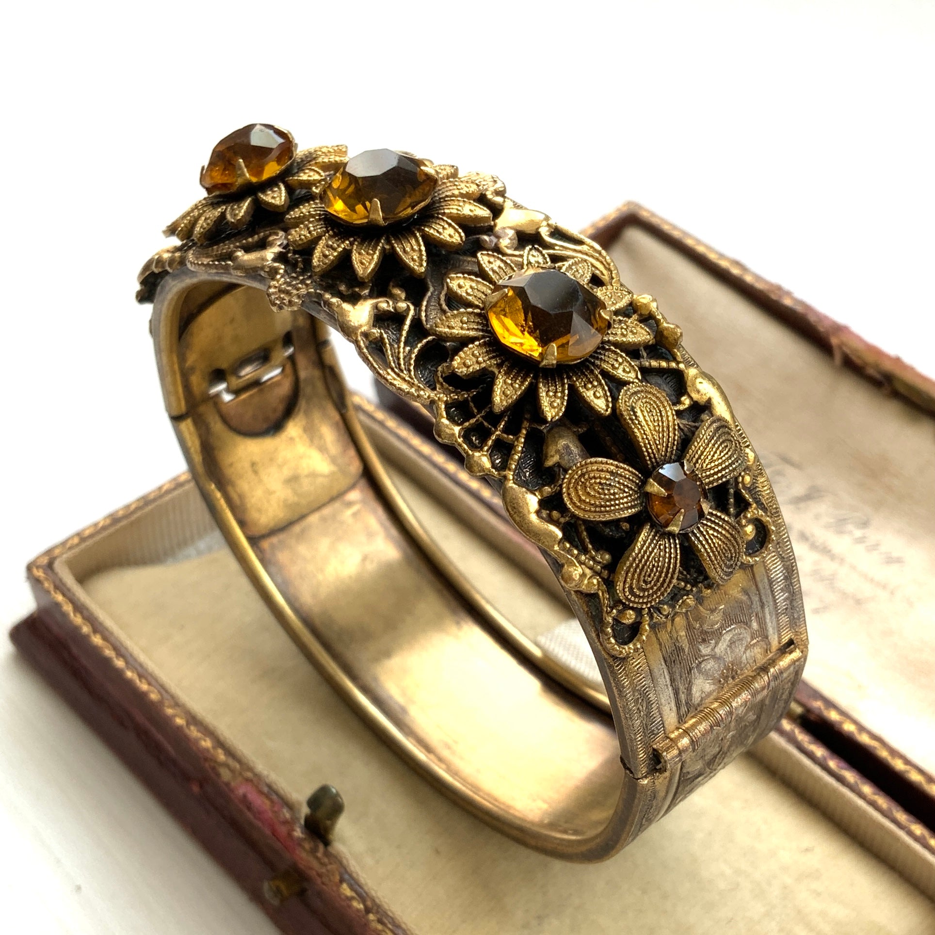 A decorative bangle featuring intricate floral motifs on gold-tone metal, with a filigree inspired design.  ゴールドトーンのメタルに、フィリグリー風の繊細な花模様があしらわれたデコラティブなバングル。