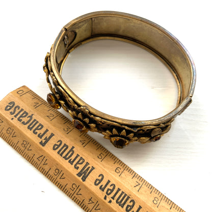 A decorative bangle featuring intricate floral motifs on gold-tone metal, with a filigree inspired design.  ゴールドトーンのメタルに、フィリグリー風の繊細な花模様があしらわれたデコラティブなバングル。