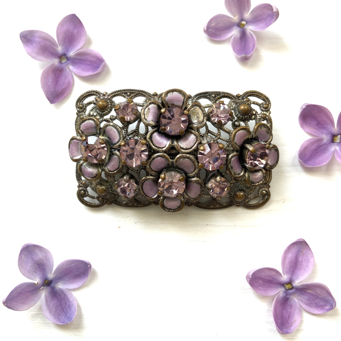 A brooch featuring a lilac-colorued flower and large matching rhinestones set on a rectangular base made of delicate filigree. 繊細なフィリグリーでできた長方形のベースに、ライラックカラーのお花と大粒の同色のラインストーンが光るブローチ。