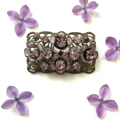 A brooch featuring a lilac-colorued flower and large matching rhinestones set on a rectangular base made of delicate filigree. 繊細なフィリグリーでできた長方形のベースに、ライラックカラーのお花と大粒の同色のラインストーンが光るブローチ。