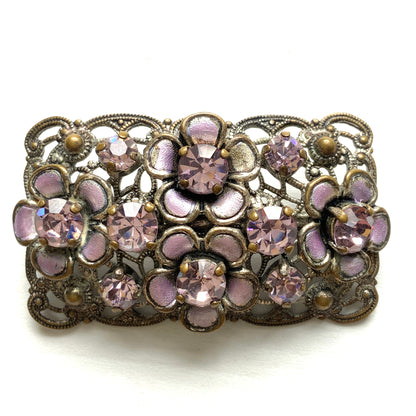 A brooch featuring a lilac-colorued flower and large matching rhinestones set on a rectangular base made of delicate filigree. 繊細なフィリグリーでできた長方形のベースに、ライラックカラーのお花と大粒の同色のラインストーンが光るブローチ。