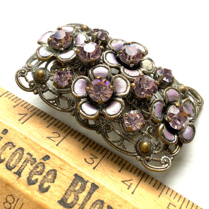 A brooch featuring a lilac-colorued flower and large matching rhinestones set on a rectangular base made of delicate filigree. 繊細なフィリグリーでできた長方形のベースに、ライラックカラーのお花と大粒の同色のラインストーンが光るブローチ。