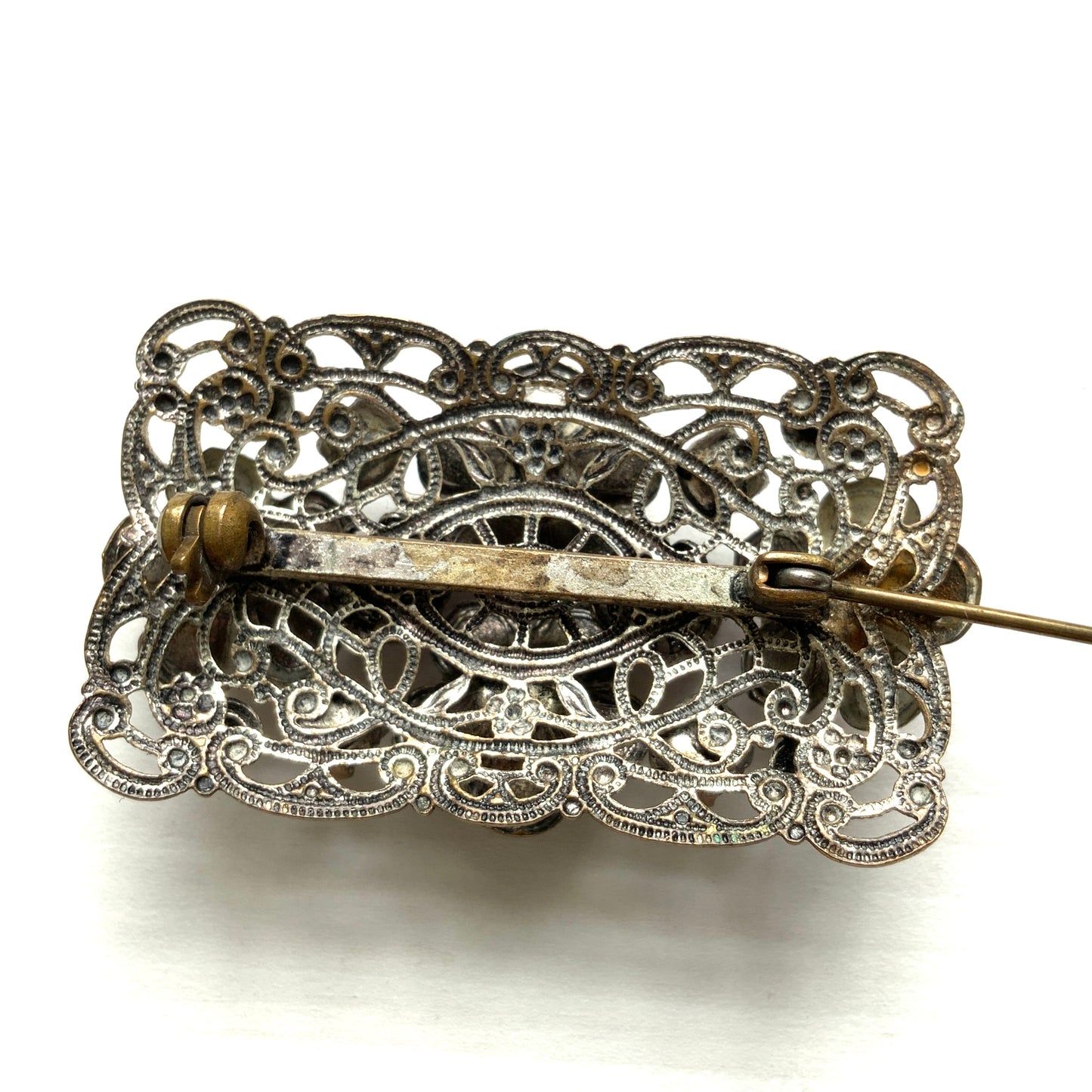 A brooch featuring a lilac-colorued flower and large matching rhinestones set on a rectangular base made of delicate filigree. 繊細なフィリグリーでできた長方形のベースに、ライラックカラーのお花と大粒の同色のラインストーンが光るブローチ。