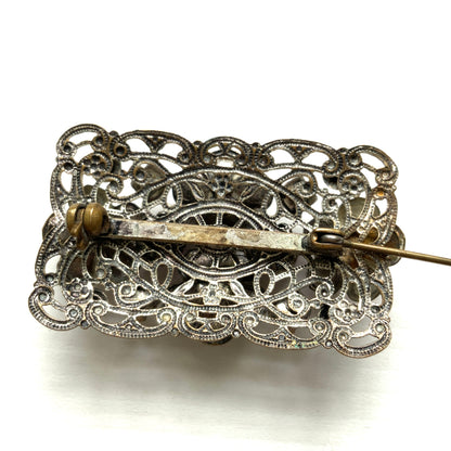 A brooch featuring a lilac-colorued flower and large matching rhinestones set on a rectangular base made of delicate filigree. 繊細なフィリグリーでできた長方形のベースに、ライラックカラーのお花と大粒の同色のラインストーンが光るブローチ。