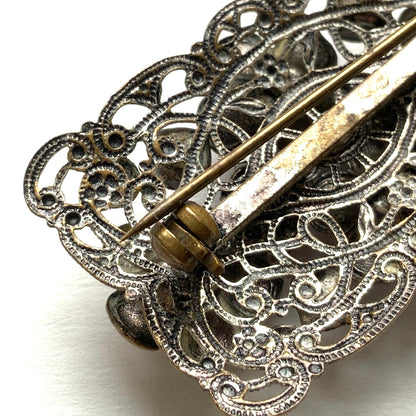 A brooch featuring a lilac-colorued flower and large matching rhinestones set on a rectangular base made of delicate filigree. 繊細なフィリグリーでできた長方形のベースに、ライラックカラーのお花と大粒の同色のラインストーンが光るブローチ。