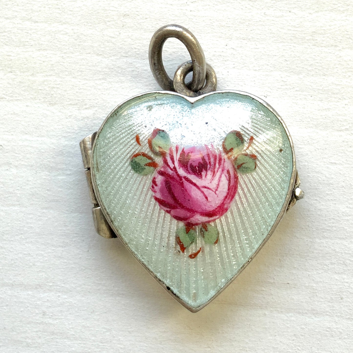 A silver heart locket featuring white guilloché enamel with a very subtle pale mint-green undertone, decorated with a rose motif.  ごく淡いミントグリーンのシェードを含んだホワイトギヨシェエナメルに、薔薇のモチーフが描かれたシルバーのハートロケット。