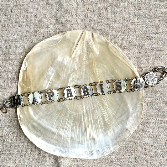 A bracelet featuring a mother of pearl tablet with the word "PARIS" and silver-tone charms of iconic Paris landmarks. マザーオブパールのタブレットに、PARISの文字とパリの観光名所のチャームがのったブレスレット。 