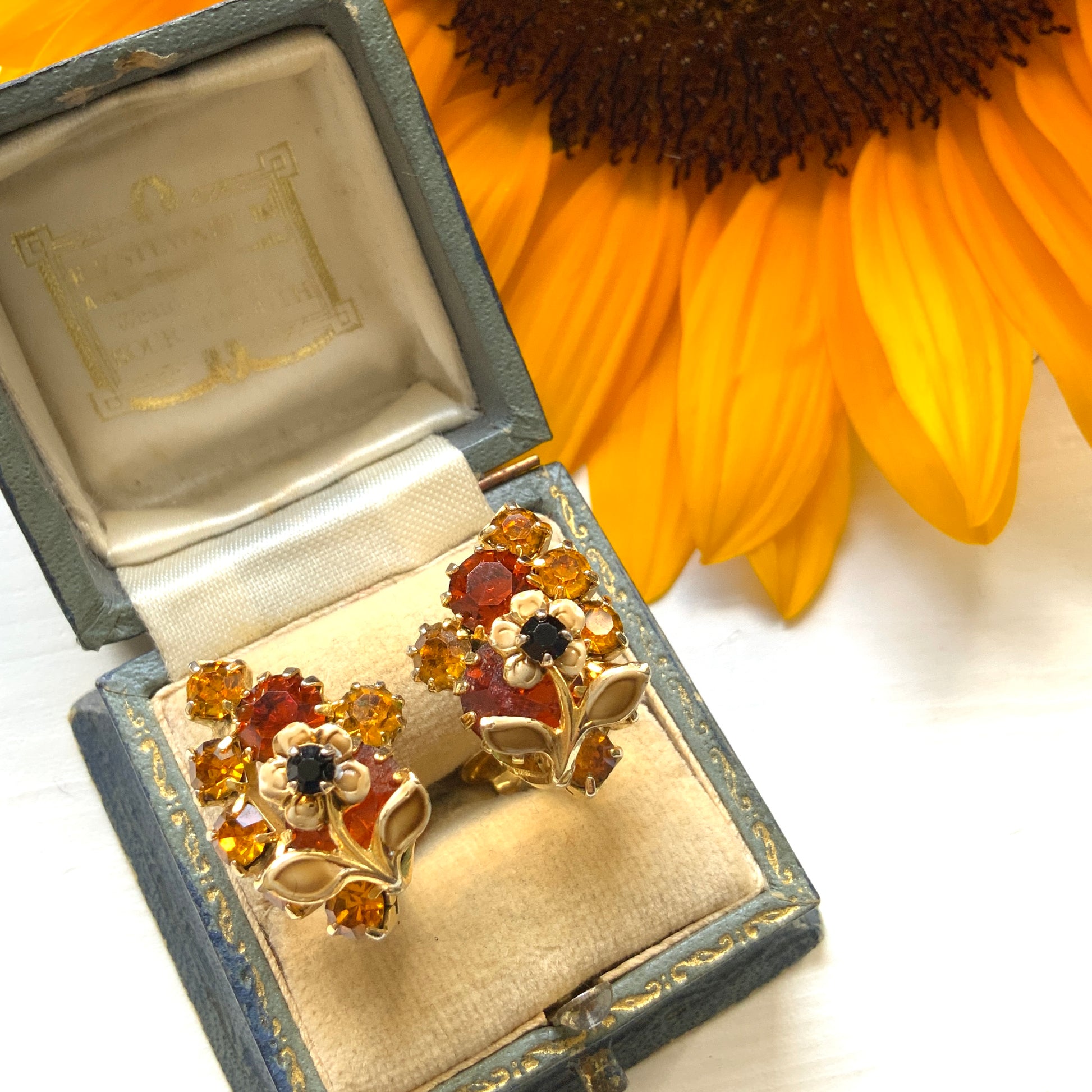 Clip-on earrings featuring vibrant orange and amber rhinestones, adorned with a petite beige enamel flower.  小さなベージュのエナメルフラワーが咲いた、アンバーラインストーンのクリップイヤリング。
