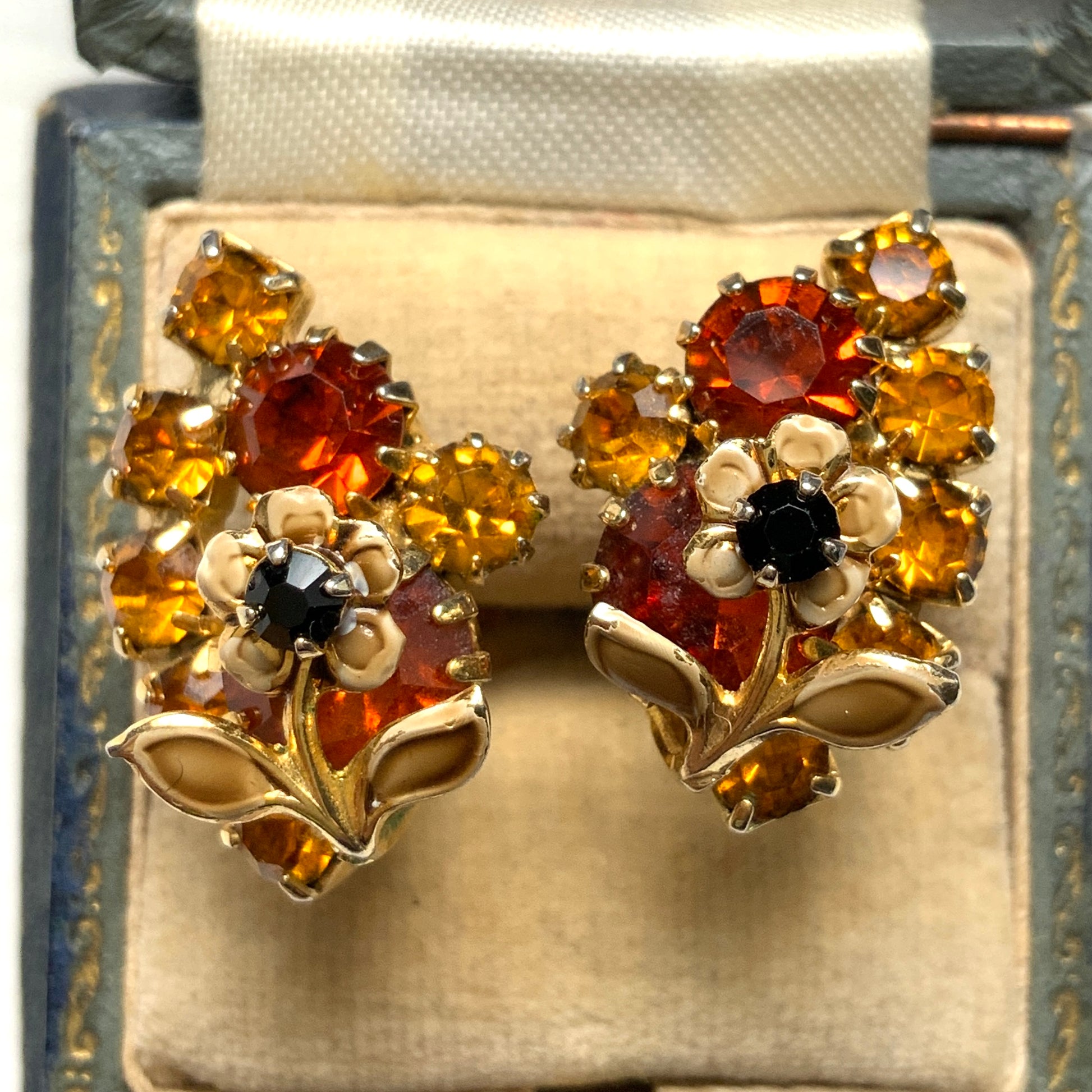 Clip-on earrings featuring vibrant orange and amber rhinestones, adorned with a petite beige enamel flower.  小さなベージュのエナメルフラワーが咲いた、アンバーラインストーンのクリップイヤリング。